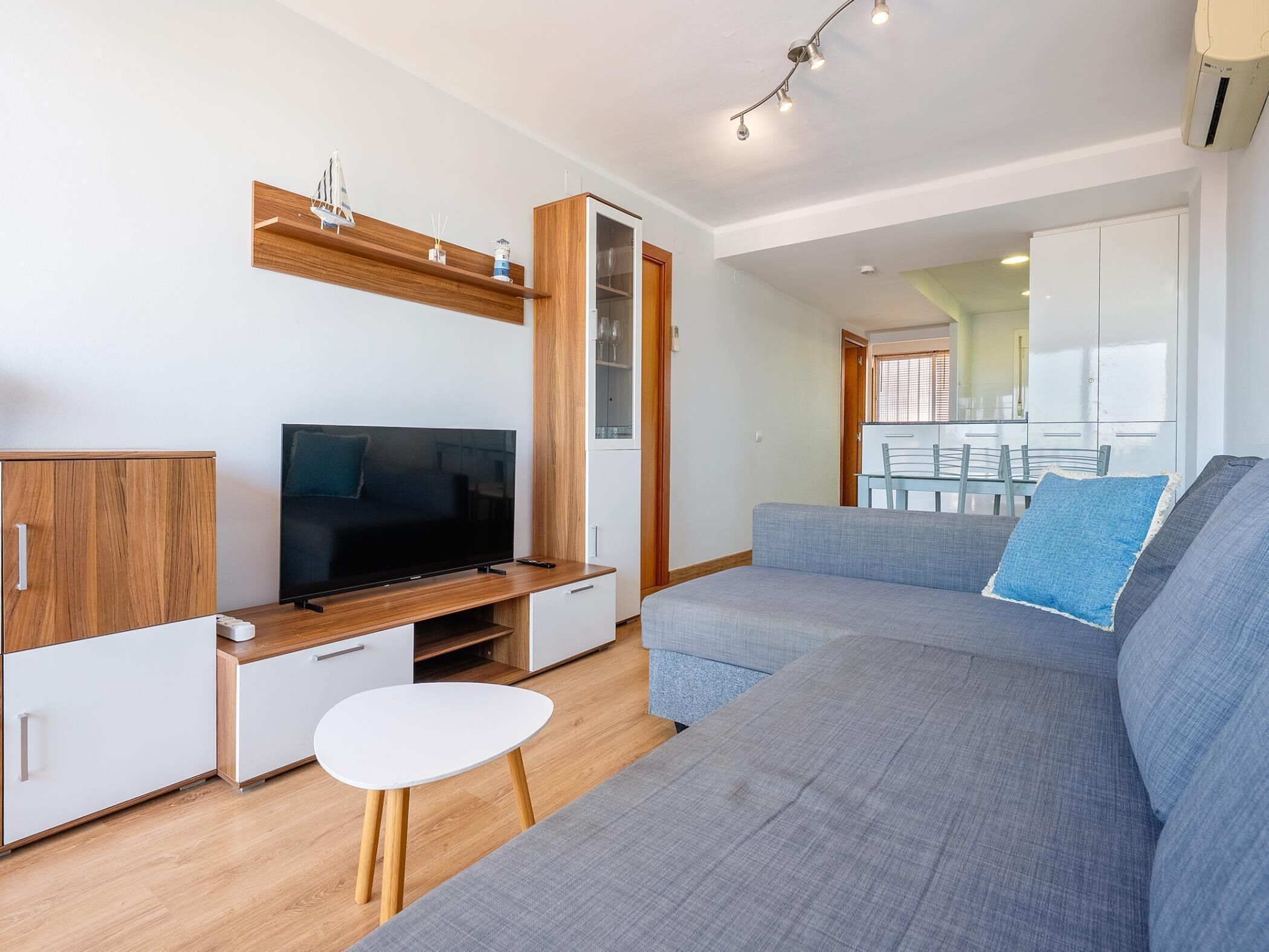 Een moderne woonkamer met een grote grijze bank, een flatscreen televisie, houten kasten en een kleine witte tafel.