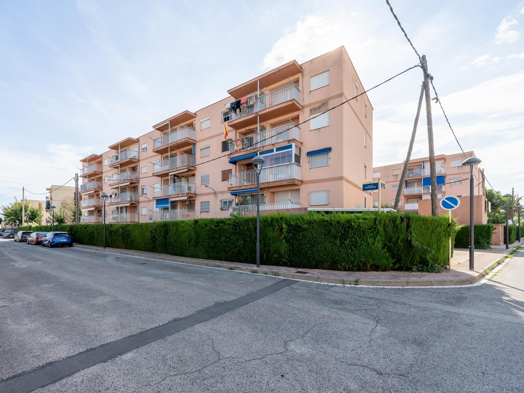 Un edificio residencial de varios pisos con balcones, cielo despejado y automóviles estacionados a lo largo de la calle.