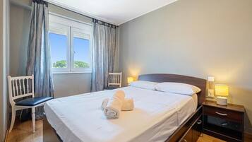 A hotel room with a bed, two bedside tables, a chair, a window with curtains, and a view of greenery outside.