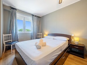 A hotel room with a bed, two bedside tables, a chair, a window with curtains, and a view of greenery outside. - Apartment with terrace 400m beach (Cambrils)