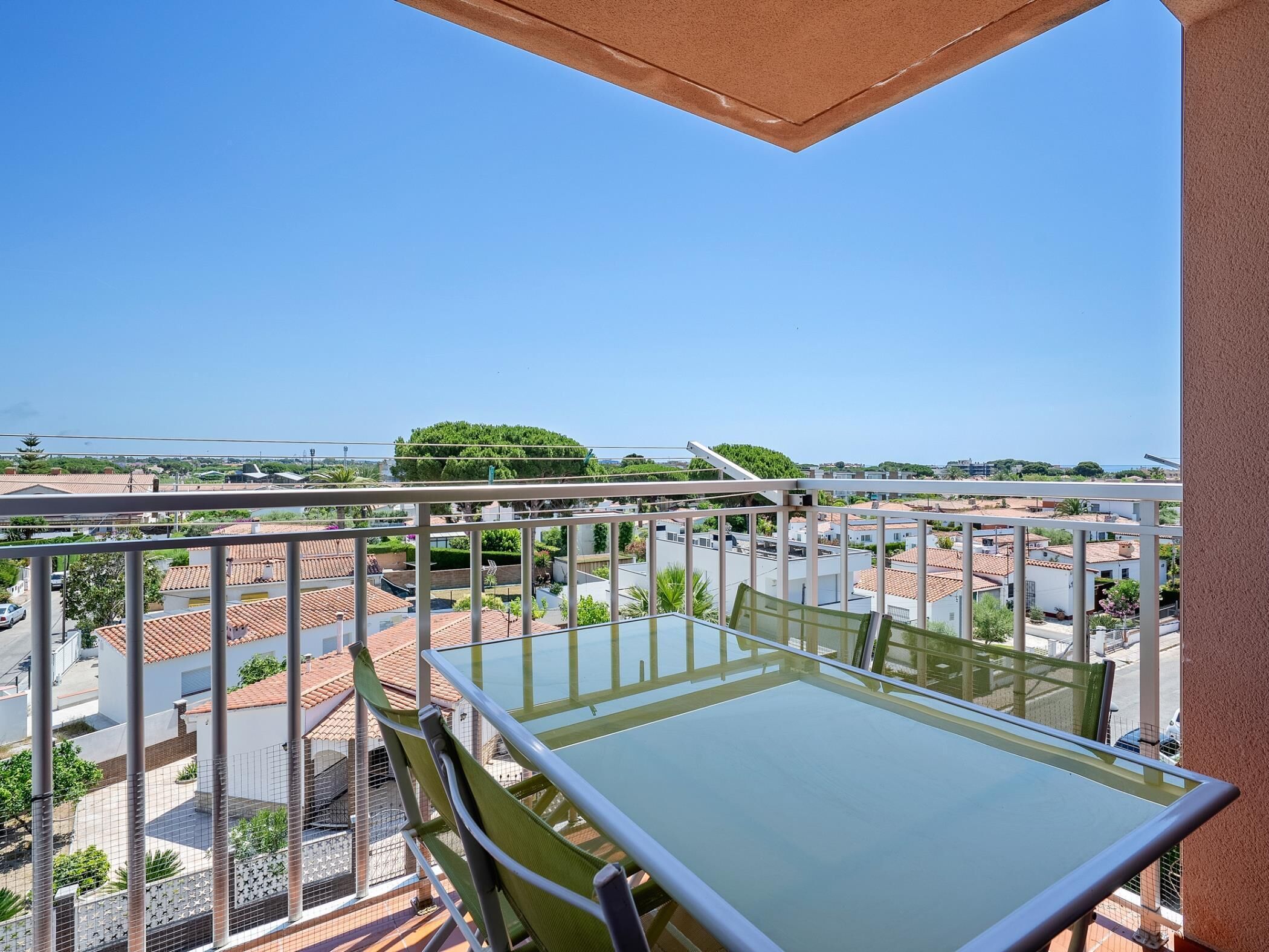A balcony with a glass table and chairs, overlooking a residential area with houses and greenery.