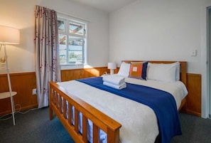 A hotel room with a wooden bed, a bedside table, a lamp, and a window with curtains. - Classic 3-Bedroom Home in Whangarei (Whangārei)