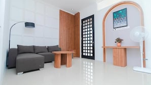 A modern living room with a sofa, a coffee table, a floor lamp, a wooden door, and a wall-mounted fan. - Cozzy Villa Ree (batu)