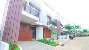 A modern two-story residential building with a brick facade, a balcony, and a landscaped front yard with various plants. - Cozzy Villa Ree (batu)