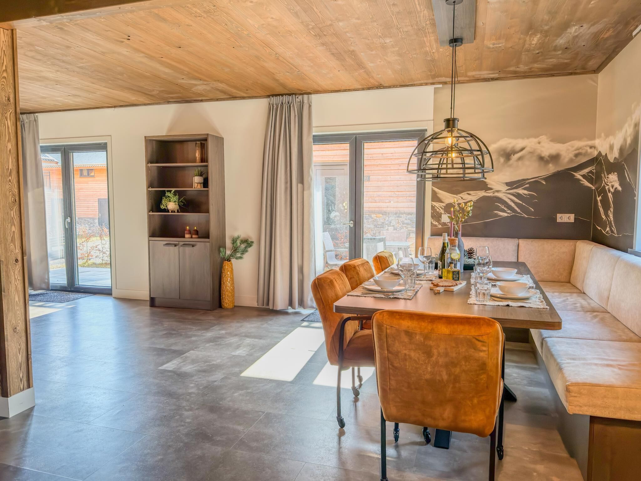 A dining area with a wooden table, chairs, and a sofa. There is a bookshelf and a large window with curtains.