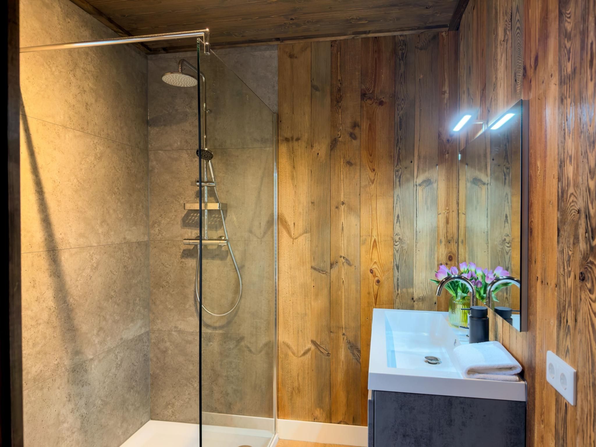 A modern bathroom with a glass-enclosed shower, wooden panel walls, a white sink with a mirror, and a vase with flowers.