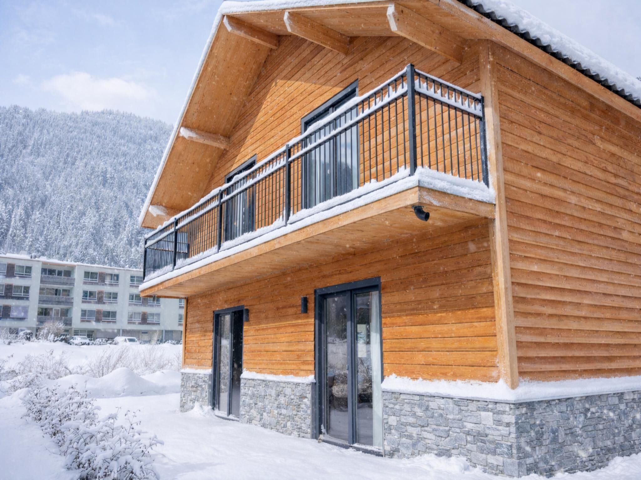 A wooden chalet with a balcony, stone foundation, and a snowy landscape.