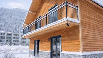 A wooden chalet with a balcony, stone foundation, and a snowy landscape.