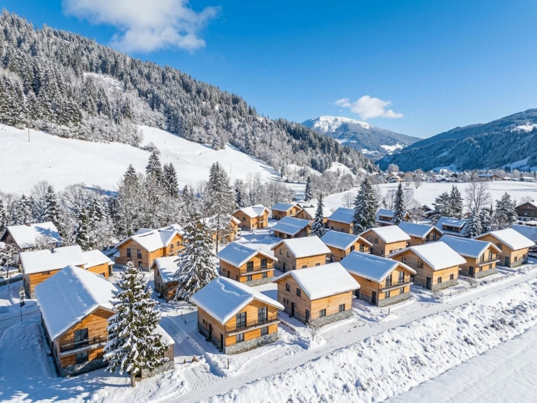 A cluster of wooden cabins with snow-covered roofs, surrounded by a snowy landscape and trees, with mountains in the background.
