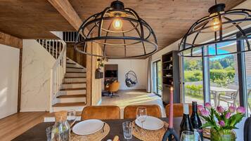 A modern dining area with a wooden table set for a meal, a hanging chair, and a view of a garden through large windows.