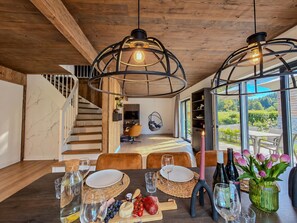 Un espace repas moderne avec une table en bois dressée pour un repas, un fauteuil suspendu et une vue sur un jardin, visible à travers de grandes fenêtres.