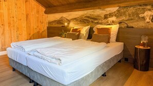 Ein Schlafzimmer mit Holzbalkendecke, einem Bett mit weißer Bettwäsche, einem Wandgemälde von einer Kuh und einem kleinen Holztisch mit Kerze.