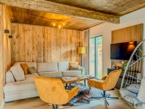 Un salon moderne avec un plafond en bois, un canapé d’angle, deux fauteuils, une table basse et un téléviseur fixé au mur.