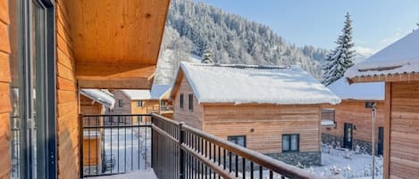 A wooden cabin with a balcony, snow-covered ground, and a mountainous backdrop.