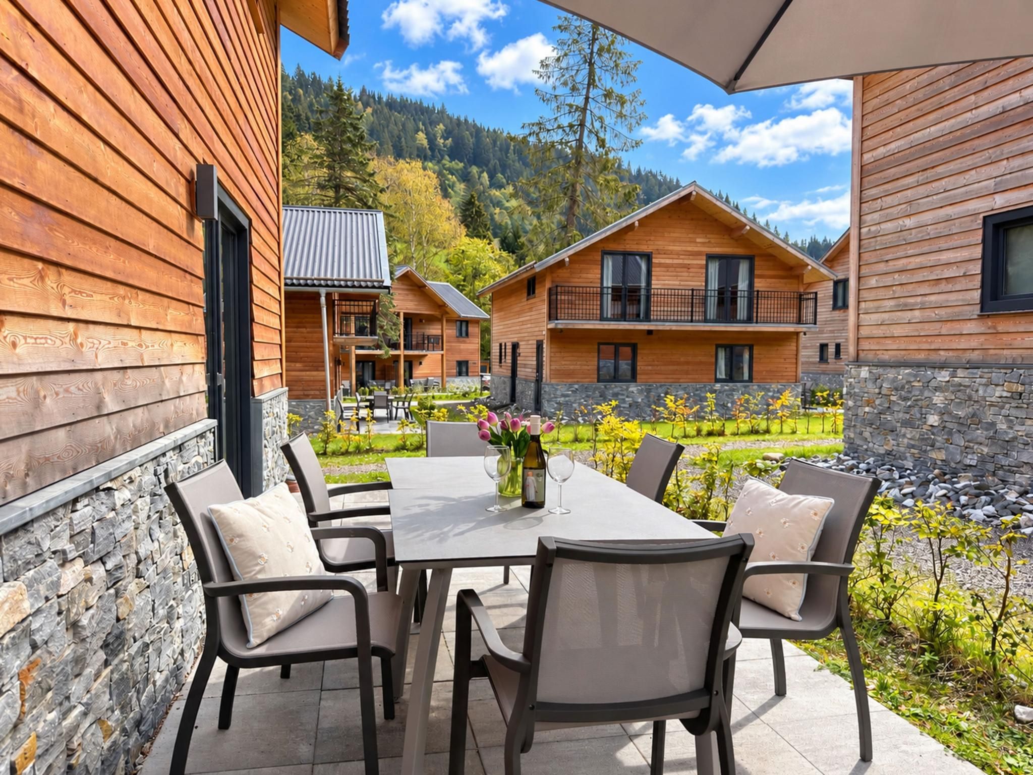 A wooden cabin with a patio table and chairs, surrounded by a stone wall and a view of a forested mountain.