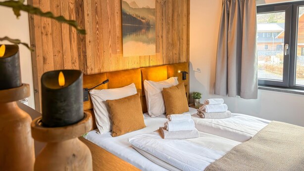Ein Hotelzimmer mit Bett, Kissen, einem Gemälde, einem Fenster mit Vorhängen und einer Vase mit Kerze.