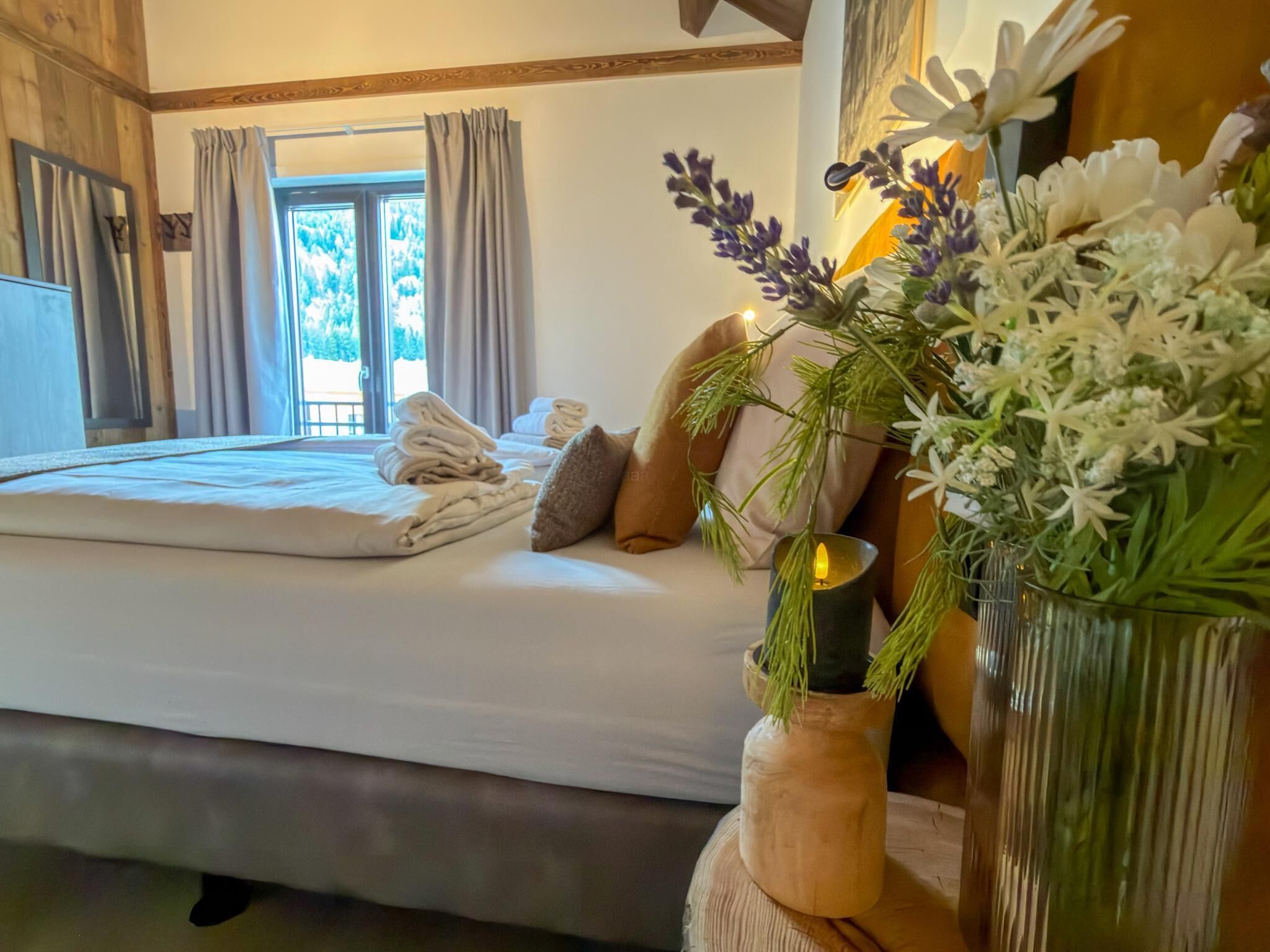 A bedroom with a bed, pillows, a vase with flowers, and a window with curtains.