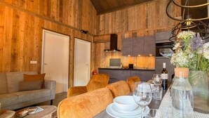 Ein gemütliches Zimmer mit Holzvertäfelung, einer Couch, einem Esstisch gedeckt mit Tellern, Gläsern und Besteck sowie einer Küchenzeile im Hintergrund.