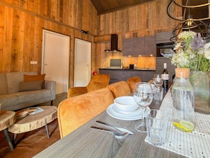 Een gezellige kamer met houten inrichting, een bank, een eettafel gedekt met borden, glazen en bestek, en een keuken in de achtergrond.
