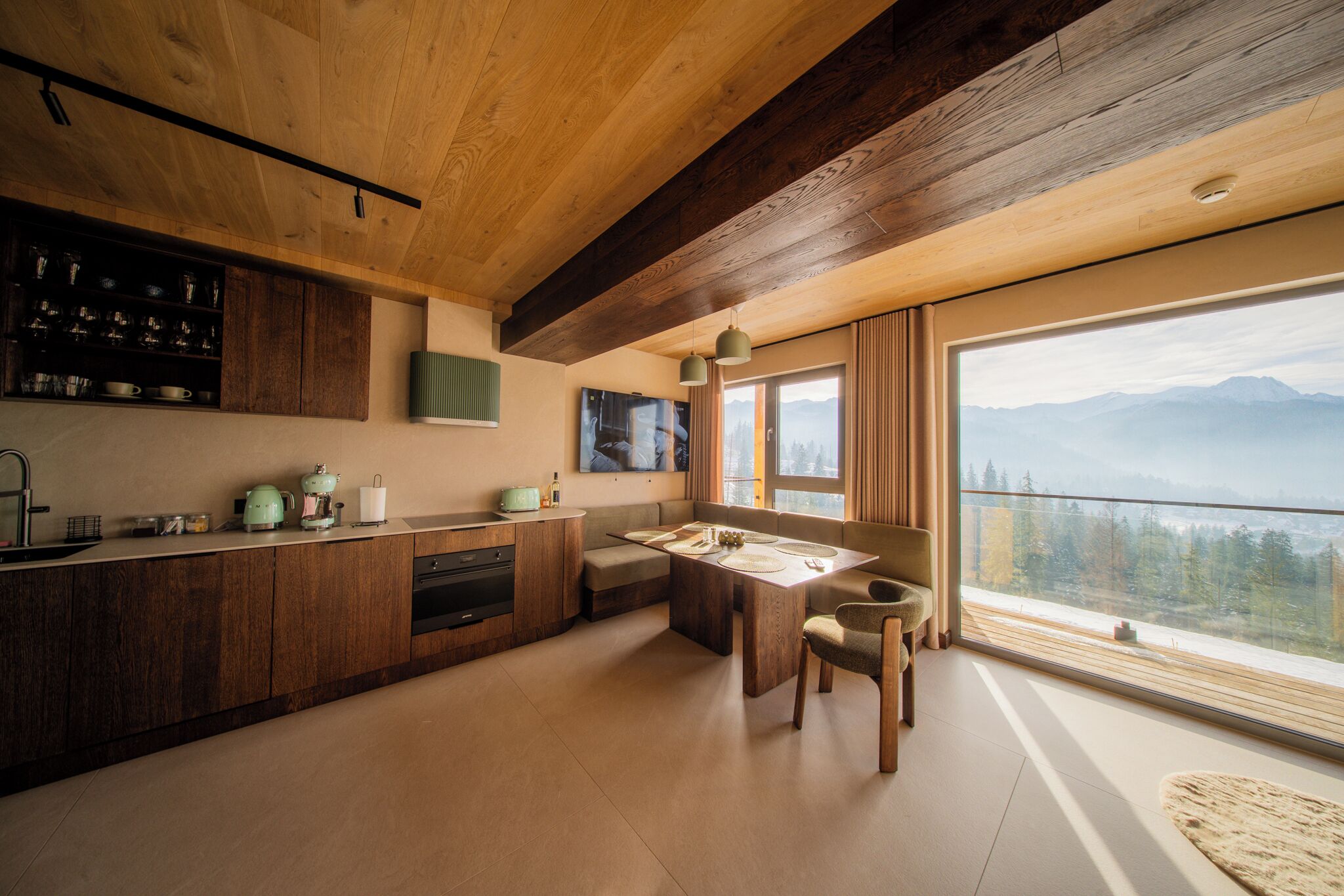 A modern kitchen with wooden cabinets, a dining area with a table and chairs, and a large window offering a mountain view.