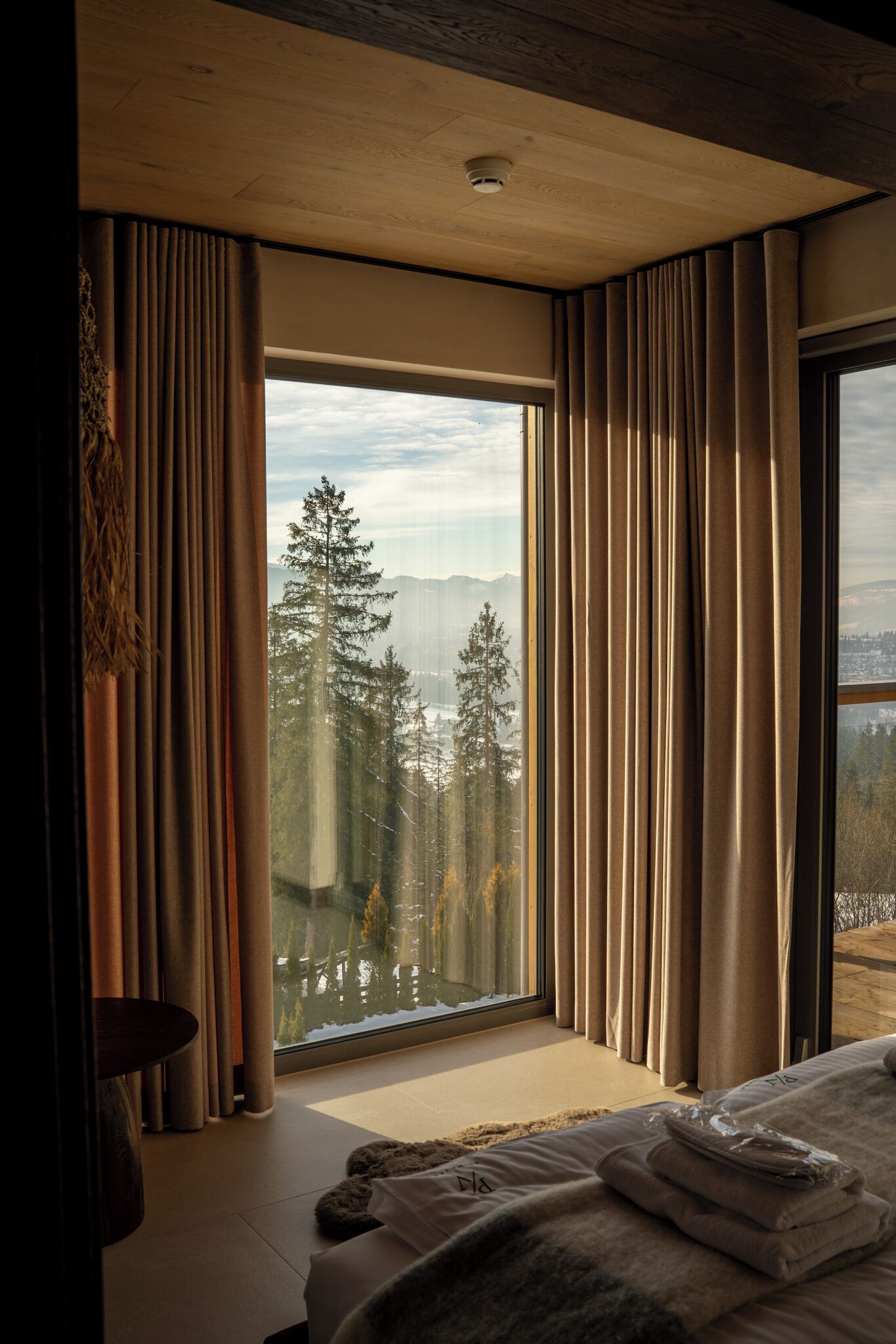 Une chambre avec une grande fenêtre, des rideaux, un lit et une vue sur les arbres et les montagnes.
