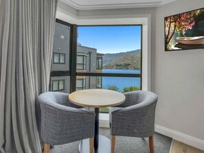 Een kamer met een groot raam met uitzicht op een meer en bergen, een ronde tafel met twee stoelen, en een schilderij aan de muur.