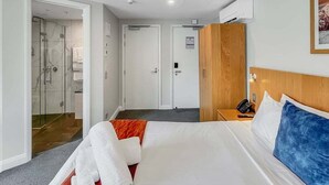 Quarto de hotel com cama, cabeceira de madeira, escrivaninha e telefone, e área de chuveiro.