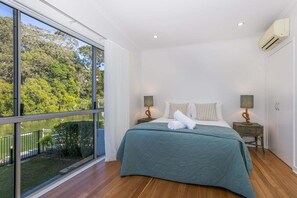A bedroom with a large window, a bed with a green bedspread, two bedside tables with lamps, and an air conditioning unit on the wall. - Luxury Waterfront Stay at Tallebudgera Creek (Gold Coast)