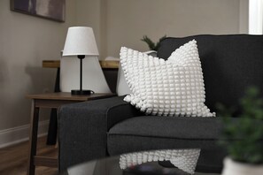 Un salon moderne avec un canapé foncé, un coussin blanc à texture particulière, une lampe sur une table d’appoint et un reflet sur une surface de verre.