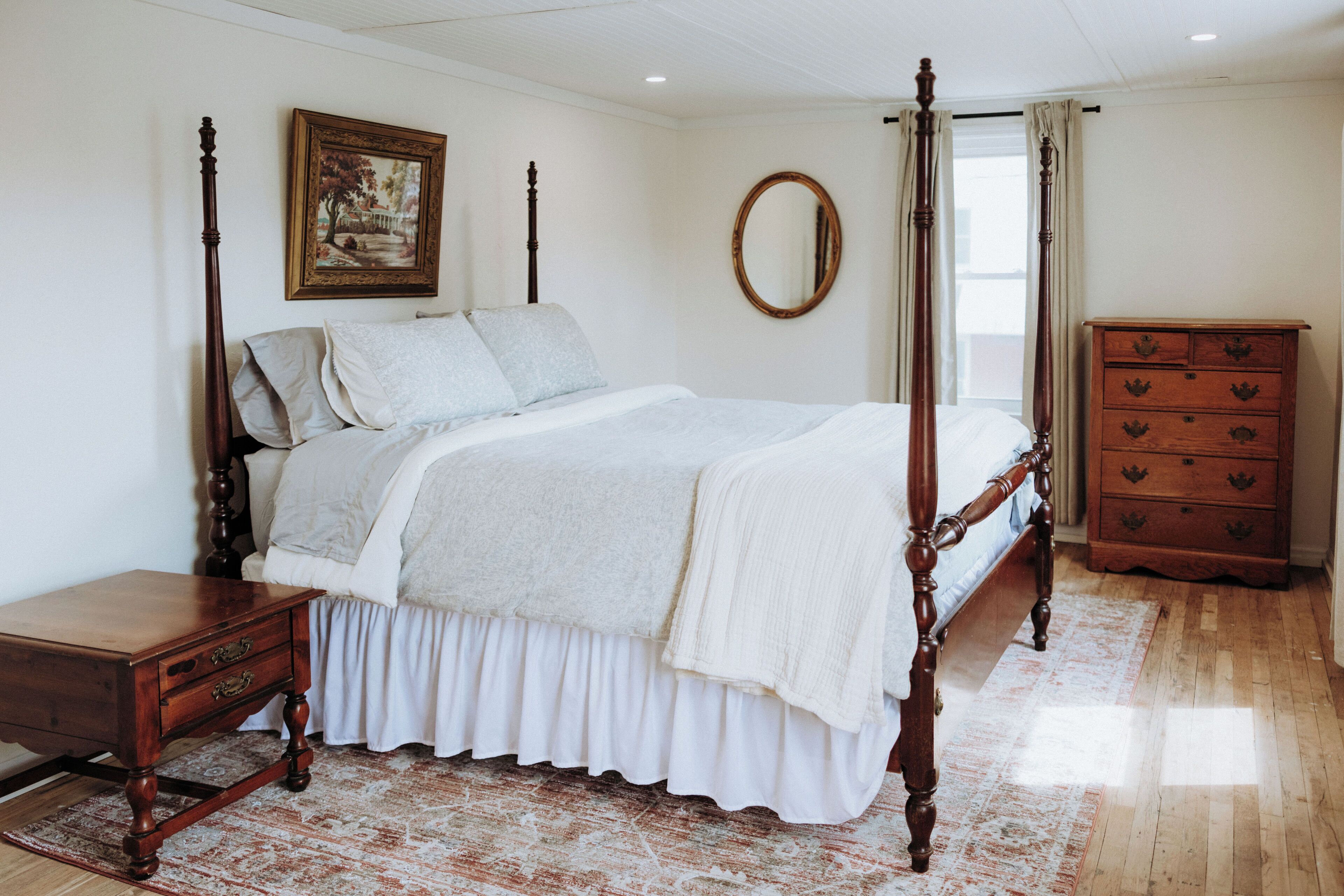 Un lit à baldaquin avec un couvre-lit blanc et des coussins assortis, une table de chevet en bois, une commode à tiroirs, un miroir rond accroché au mur et un tapis à motifs sur le sol.