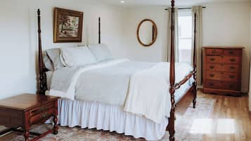 Un lit à baldaquin avec un couvre-lit blanc et des coussins assortis, une table de chevet en bois, une commode à tiroirs, un miroir rond accroché au mur et un tapis à motifs sur le sol.
