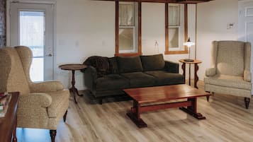 Une pièce avec un plancher en bois, un canapé vert, une table basse en bois, deux fauteuils et une petite table ronde.