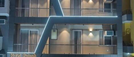 A multi-story hotel with a modern design, featuring a prominent 'Hotel Palash' sign, balconies, and a green carpeted entrance.