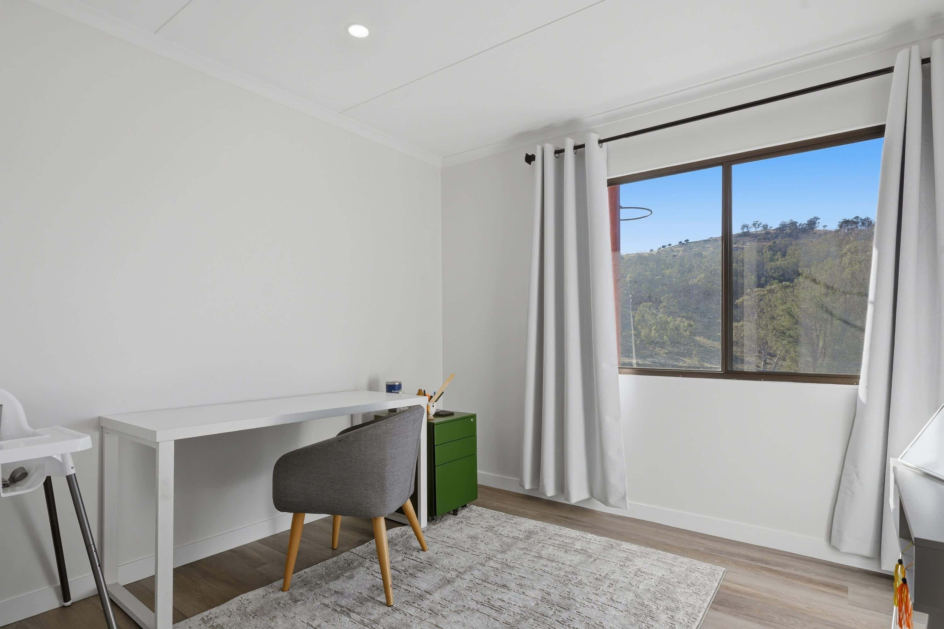 Une pièce moderne avec un bureau blanc, une chaise grise, un meuble vert et une grande fenêtre avec des rideaux.
