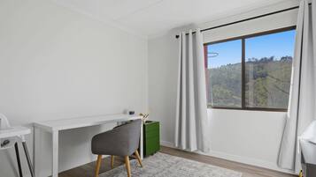 Une pièce moderne avec un bureau blanc, une chaise grise, un meuble vert et une grande fenêtre avec des rideaux.