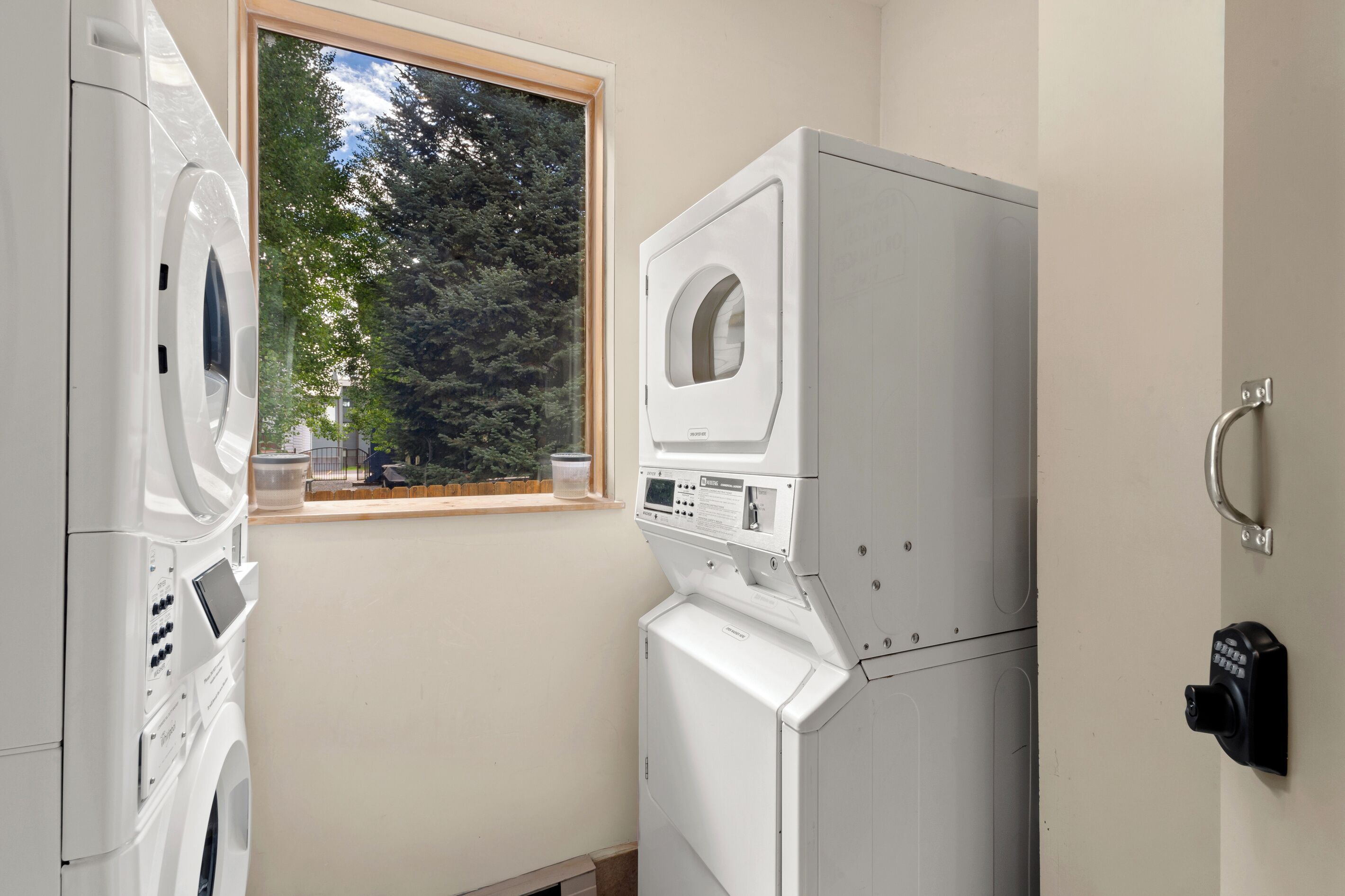 A laundry room with stacked washers and dryers, a window with a view of trees, and a wall-mounted security device.