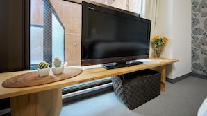 Een flatscreen televisie op een houten tv-meubel met een geweven onderzetter, een kleine cactusplant en een vaas met bloemen.