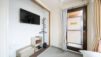 Quarto de hotel com televisão de tela plana na parede, um porta-roupas, um pequeno espaço de estar com almofada e uma porta com uma placa.