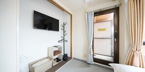 A hotel room with a flat-screen TV mounted on the wall, a coat rack, a small seating area with a cushion, and a door with a sign.