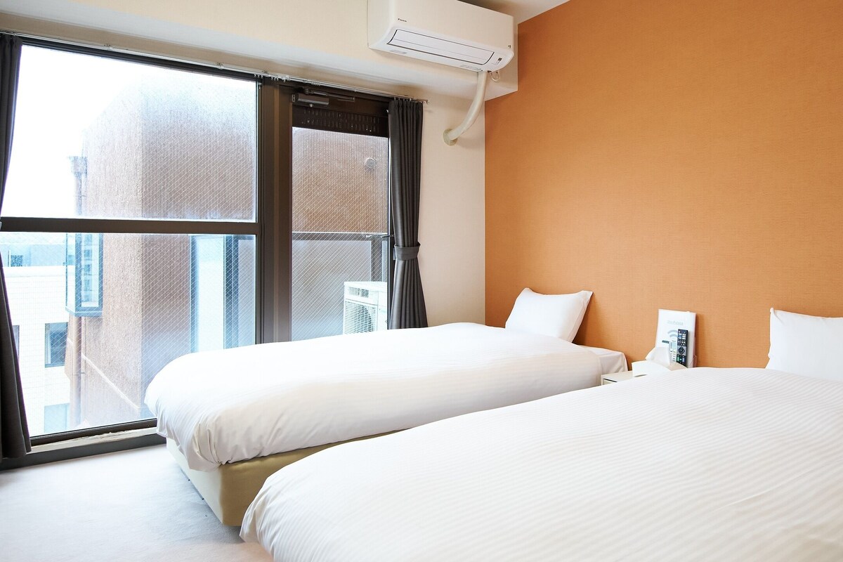 Una habitació d'hotel amb dos llits, un finestra gran amb cortines, un aparell d'aire condicionat i vistes a la ciutat.