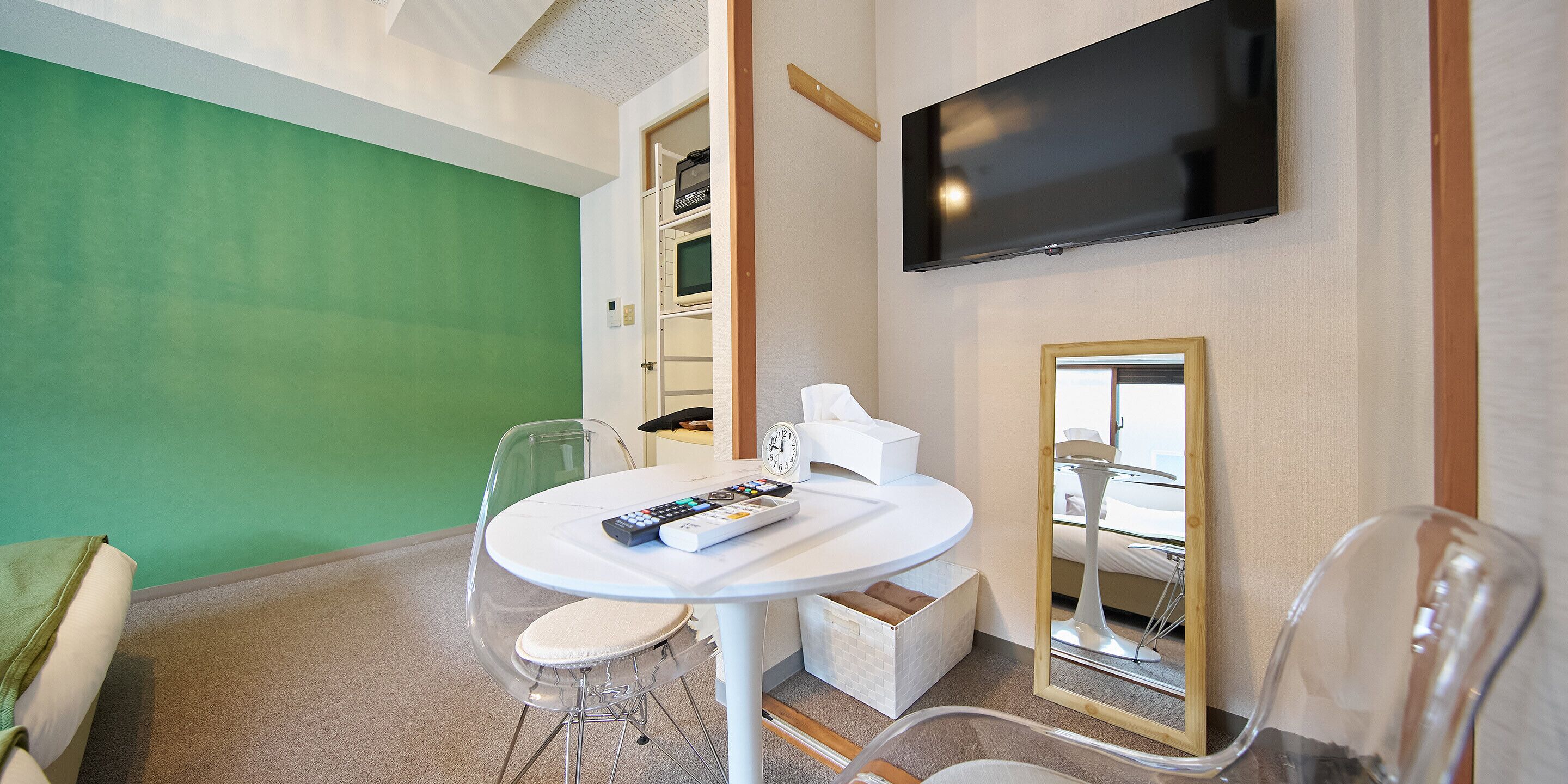 A hotel room with a round white table, a flat-screen TV mounted on the wall, a green accent wall, and a mirror with a wooden frame.