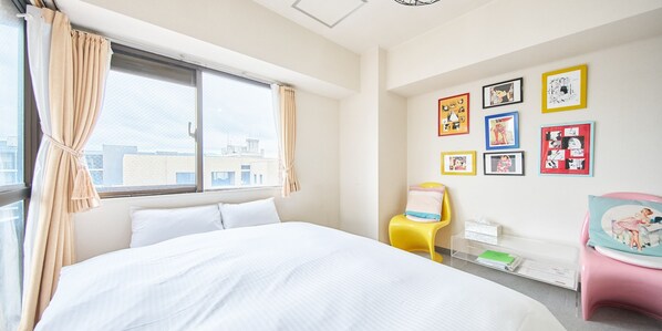A bedroom with a bed, a window with curtains, a wall with framed pictures, and a small table with a chair.