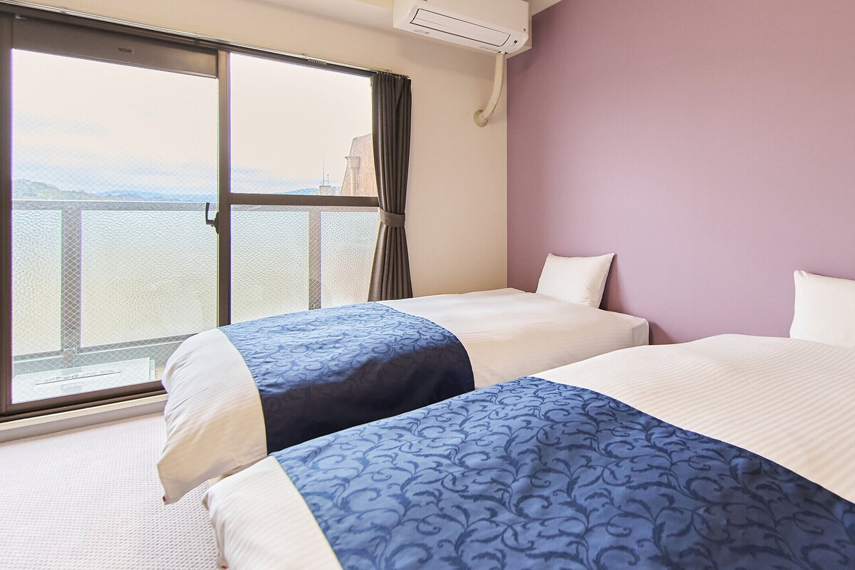 Habitació d'hotel amb dos llits, una finestra gran i un aparell d'aire condicionat.