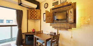 A compact room with a television mounted on the wall, a small table with a chair, a red kettle, and a clock.