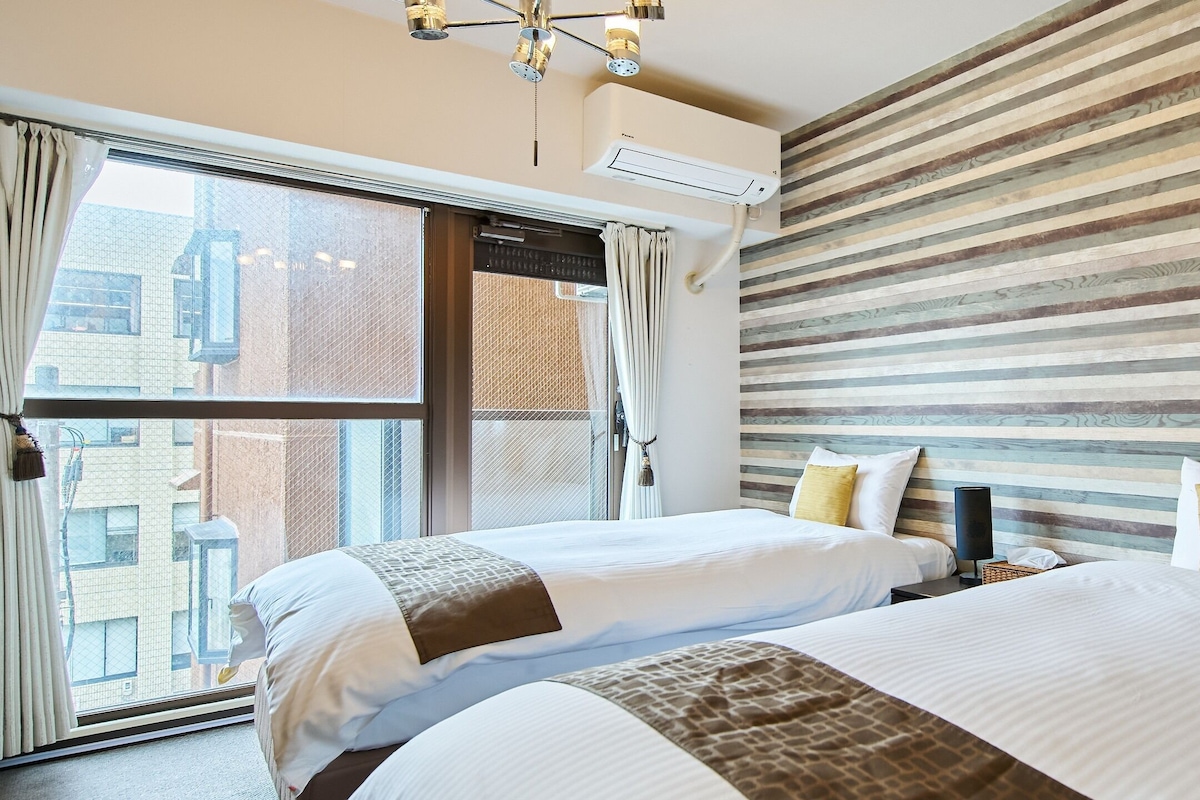 Habitació d'hotel amb dos llits, una finestra gran amb cortines, un ventilador de sostre i una paret amb ratlles.