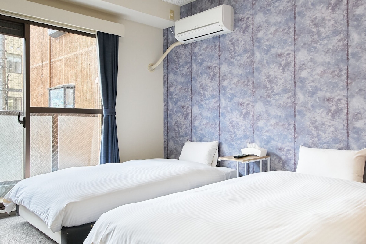 Habitació d'hotel amb dos llits, una finestra que dona a edificis i un aparell d'aire condicionat a la paret.