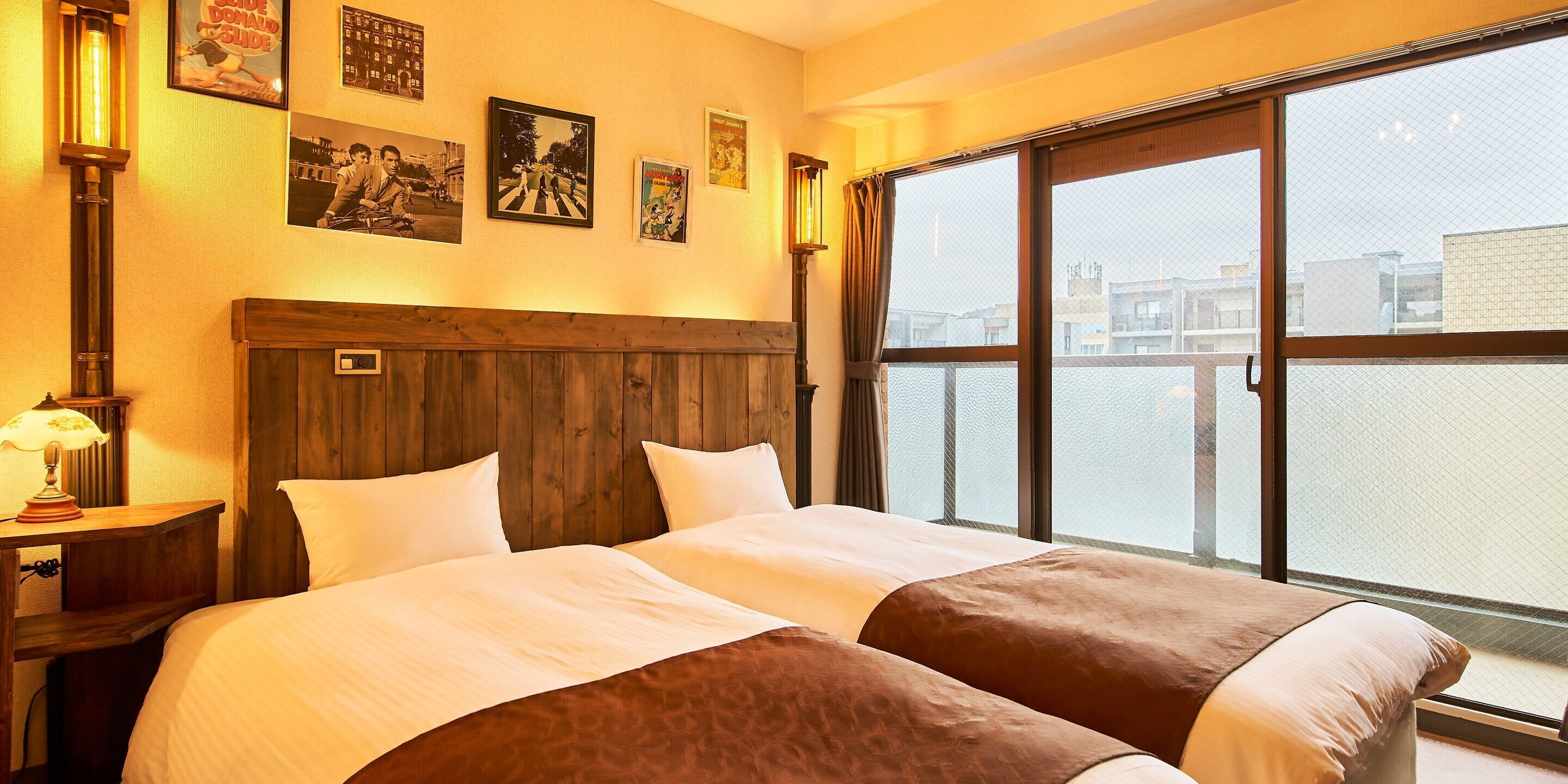 A hotel room with a large bed, a wooden headboard, a nightstand with a lamp, and a window with a view of buildings.