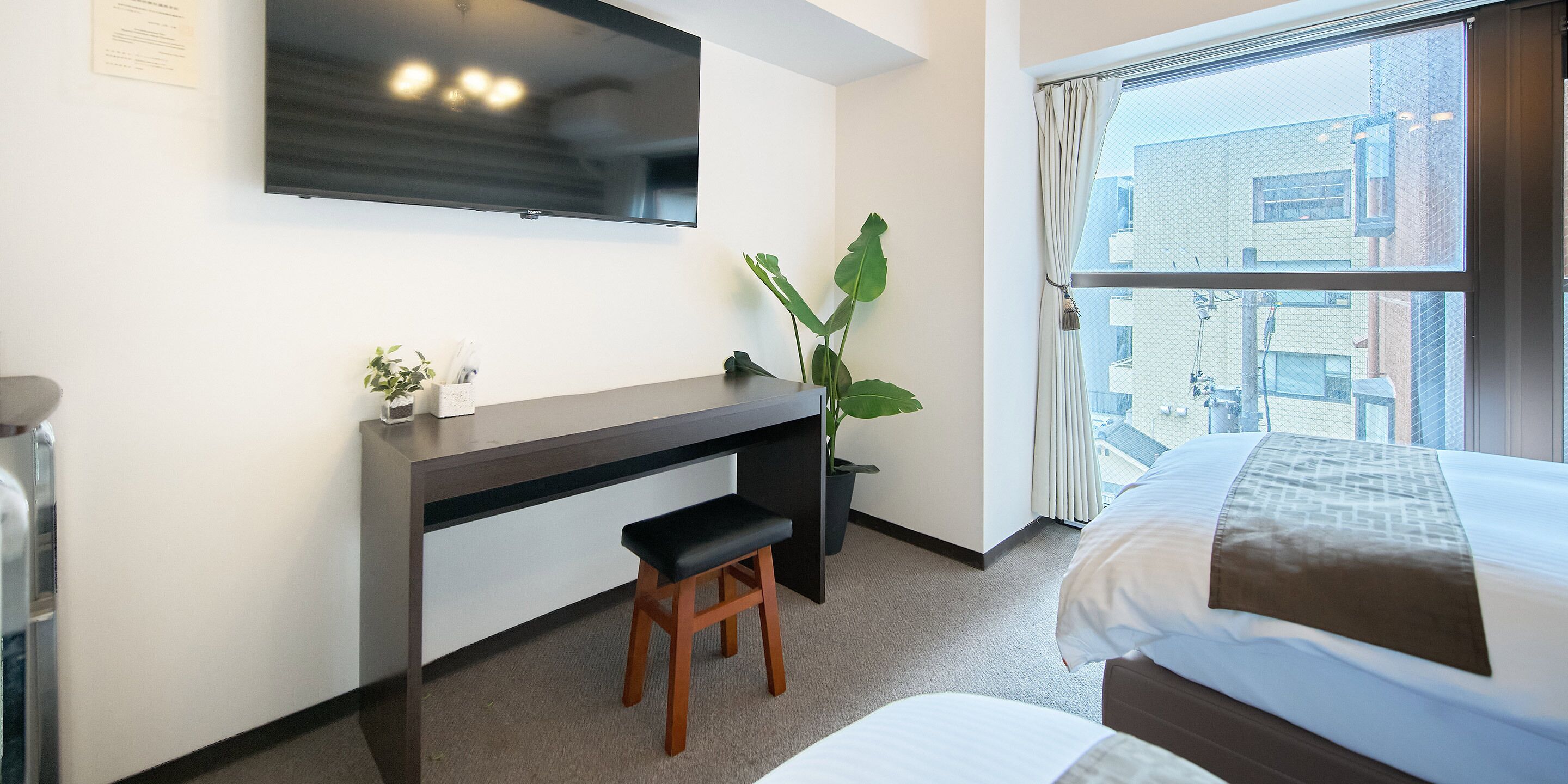 Een hotelkamer met een bed, een bureau met een stoel, een televisie en een groot raam met uitzicht op gebouwen.