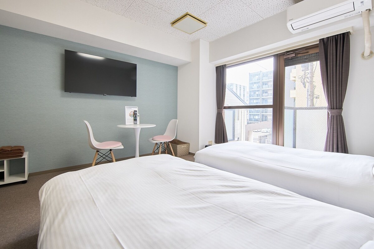 Habitació d'hotel amb dos llits, un televisió de pantalla plana penjada a la paret, una taula petita amb cadires i una finestra amb cortines.