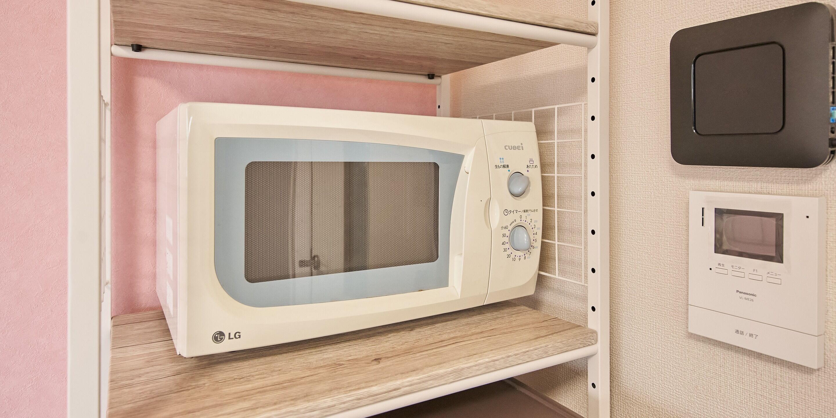 Un four à micro-ondes doté d’un écran numérique et d’un panneau de commande, installé sur une étagère dans une cuisine avec un mur rose et un plan de travail en bois.
